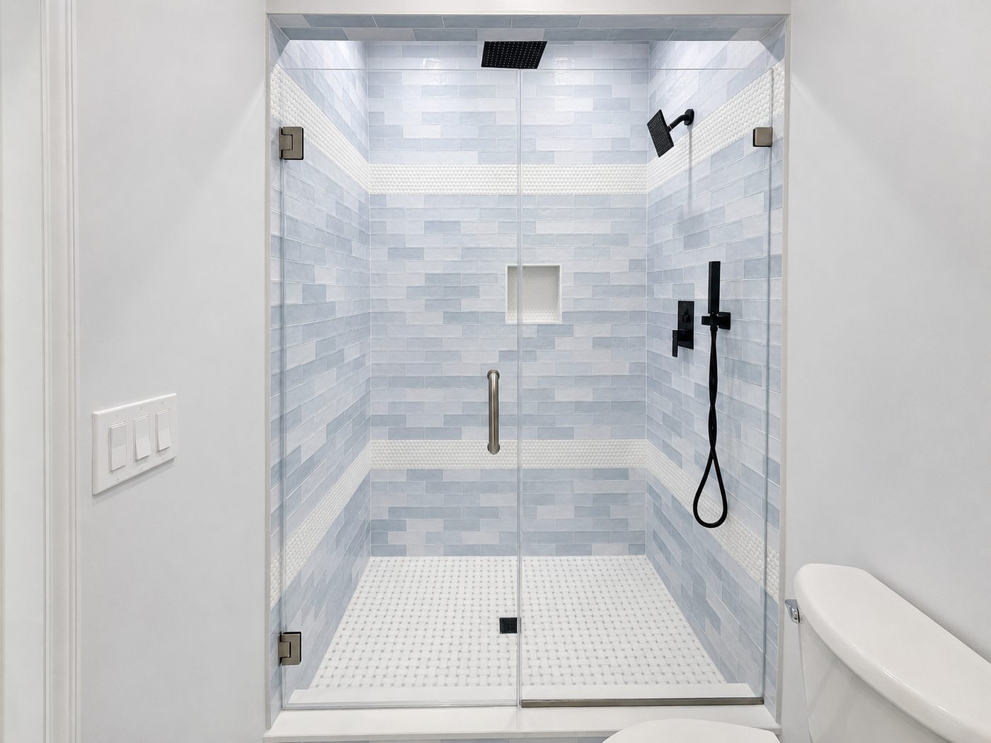 Frameless door and panel with brushed nickel hinges and pull on pale blue subway tile, matte black fixtures